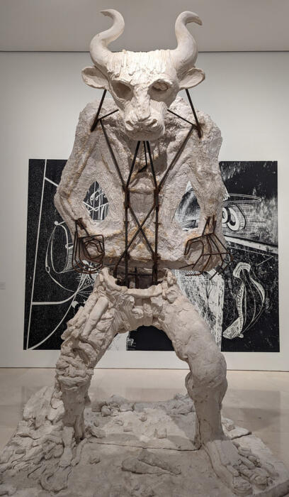 A large white sculpture of a bull-headed figure with exposed metal framework, set against a contrasting black and white art backdrop.